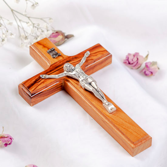 A handcrafted olive wood Latin crucifix with a figure of Jesus Christ on the cross, set against a white background with flowers.