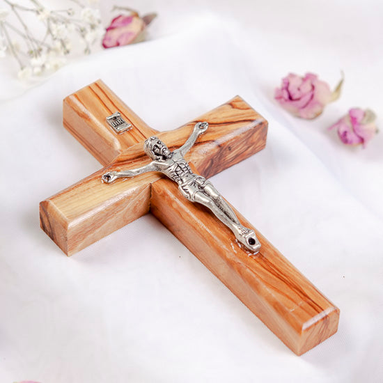 A handmade Latin crucifix made from Bethlehem olive wood, featuring silver figure of Jesus Christ, laid on white background with flowers.