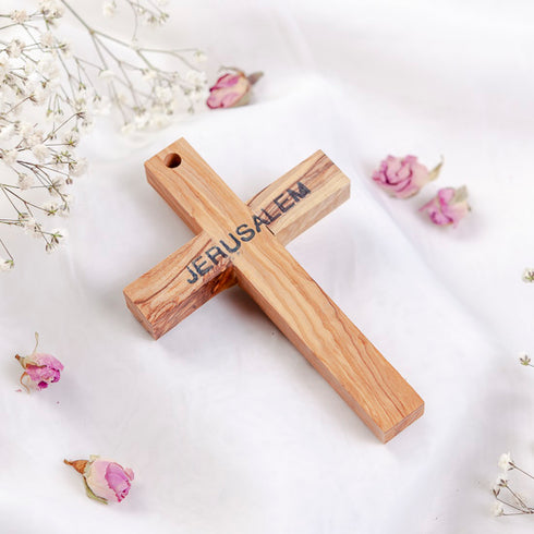 Back of a small olive wood Latin cross from Holy Land with word "Jerusalem" written, laid on white surface with rose petals.
