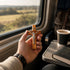 Hand holding a wooden crucifix near a window with a view of a landscape, a cup of coffee, and a book on a train or bus seat.
