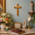 Decorative table with religious items including a cross, statue, candles, and flowers against a light blue wall.
