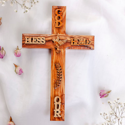 Wall-hanging olive wood Latin cross engraved with a dove and "GOD BLESS OUR HOME", on a white backdrop with rose petals.