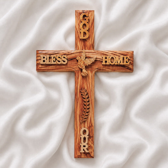 Wall-hanging olive wood Latin cross engraved with a dove and "GOD BLESS OUR HOME", on a white backdrop with rose petals.