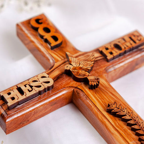 Latin olive wood cross hand-crafted and engraved with “God Bless Our Home” and a dove, laid on white background.