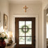 Decorative cross above a wooden door with a wreath, lamp, and flowers on a table.