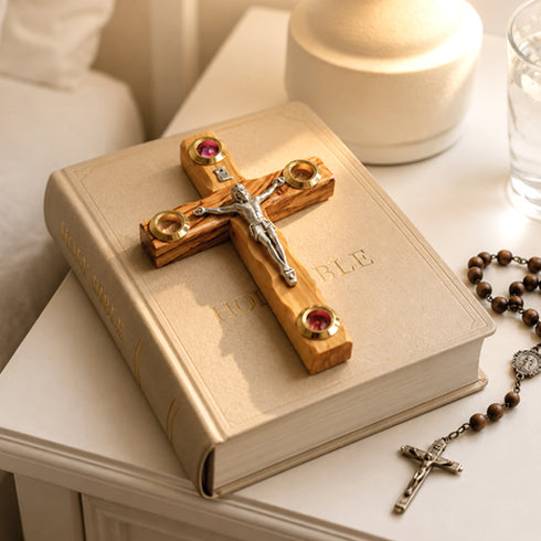 Wooden cross with red stones on an open Bible, surrounded by a rosary and a candle.