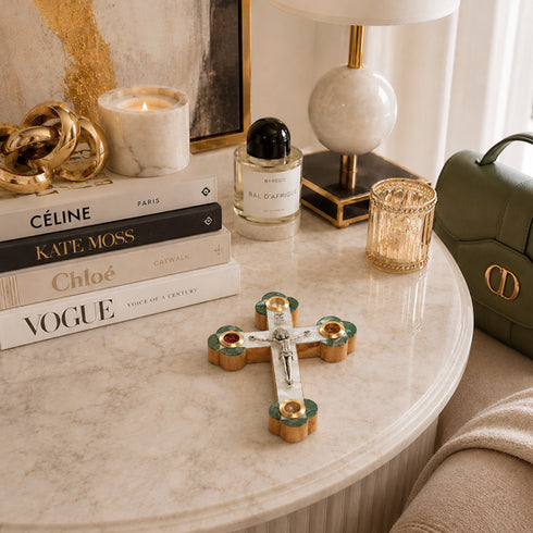 Decorative cross on a marble table with books, candles, and a lamp in the background.