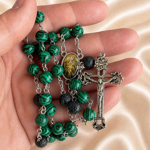 A close-up of a rosary in a hand, with green marbled and black lava stone beads, an oval medallion, and a silver-tone crucifix.