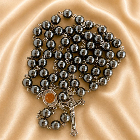 A dark gray beaded rosary with a silver-toned crucifix and centerpiece, arranged on a draped beige satin fabric background.
