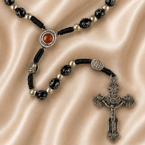 Close-up of black bead rosary with Holy Soil centerpiece, Jerusalem Cross medal, and ornate silver crucifix on satin fabric
