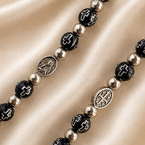 Close-up of black rosary beads with silver spacers, Virgin Mary and Jerusalem Cross medals on satin fabric background