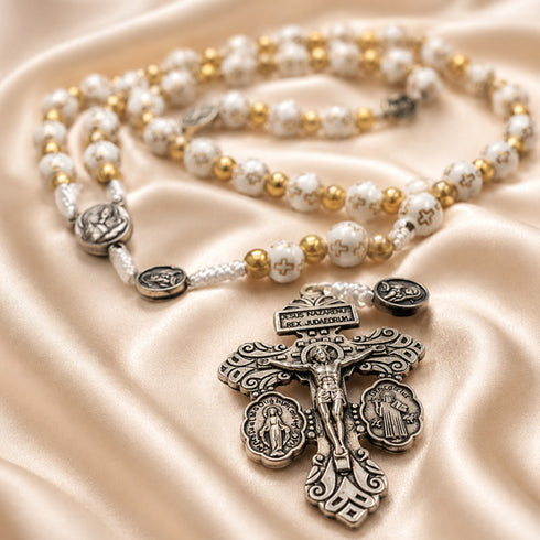 White and gold rosary with cross-pattern beads and ornate silver crucifix on a champagne satin fabric background.