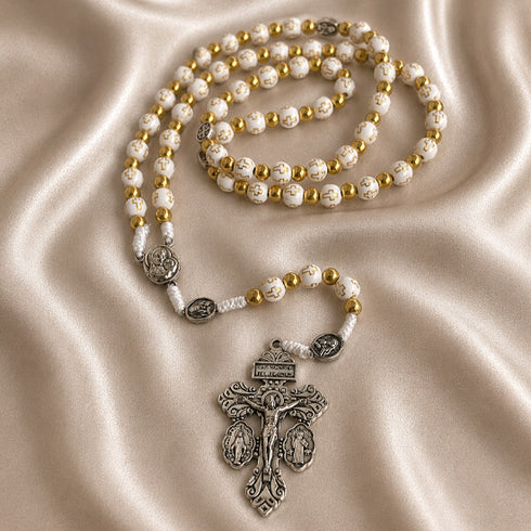 White and gold rosary with cross-pattern beads and ornate silver crucifix on a champagne satin fabric background.