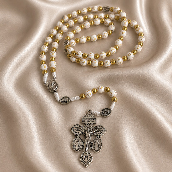 White and gold rosary with cross-pattern beads and ornate silver crucifix on a champagne satin fabric background.