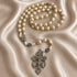 White and gold rosary with cross-pattern beads and ornate silver crucifix on a champagne satin fabric background.