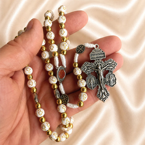 Hand holding a white and gold rosary with cross-pattern beads, a Jerusalem soil centerpiece, devotional medals, and a silver crucifix