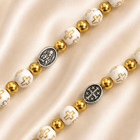 Close-up of white and gold rosary beads with Virgin Mary and Jerusalem Cross medals on a champagne satin background