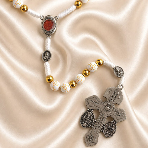 White rosary with Holy Soil centerpiece, devotional medals, and silver crucifix on a satin fabric background