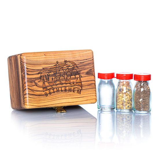 A handcrafted olive wood box against white background, with a design that includes a Bethlehem Star and panorama and the word 'Bethlehem' on the lid, accompanied by three glass vials with red caps next to it, containing holy water, incense, and holy soil.