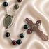 A rosary with dark green, black, and burgundy beads, a silver-tone medallion, and cross on a beige satin fabric.