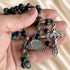 A close-up of a hand holding a beaded rosary with dark green and brown stone beads, a silver medal, and a silver crucifix.