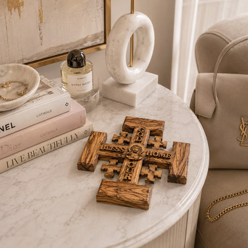 Wooden cross on a marble table with books, a bottle, and a lamp in the background.