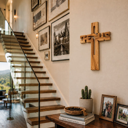 Wooden cross on a wall above a staircase with framed pictures and a potted plant.