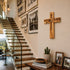Wooden cross on a wall above a staircase with framed pictures and a potted plant.