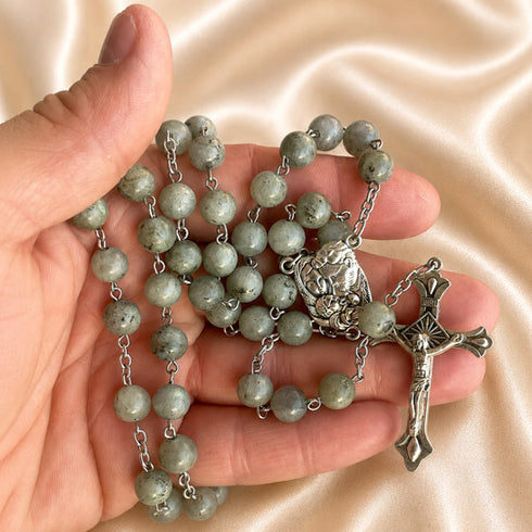 A hand holding a rosary with green stone beads, a silver-tone crucifix, a medal, against a beige background.