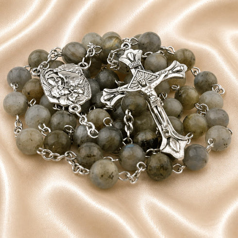 A rosary with gray and green labradorite beads, a crucifix, and a small medal pendant, on a beige satin fabric background.