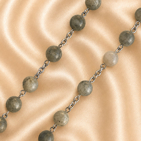 A beaded necklace with round gray stone beads and silver chain links, laid diagonally on a beige satin fabric background.