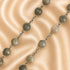 A beaded necklace with round gray stone beads and silver chain links, laid diagonally on a beige satin fabric background.