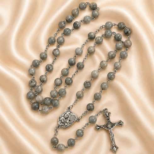 A rosary with gray stone beads and a silver-tone crucifix and centerpiece, arranged on draped satin fabric.