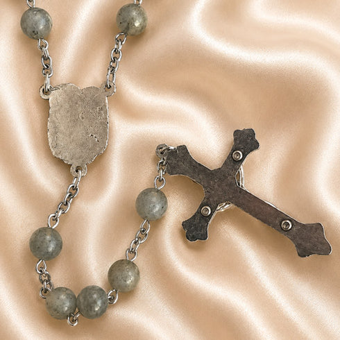 A rosary with round gray stone beads, a silver-tone centerpiece, and a metal crucifix, on a satin beige fabric background.