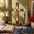 Religious decor with a cross, statue of a Virgin Mary, open book, rosary, and candle on a table.