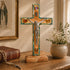 Decorative cross with crucifix on a wooden base, placed on a table with a book and vase in the background.