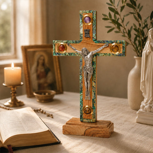 Decorative cross with Jesus Christ on a wooden base on a table with a candle, book, and statue in the background.