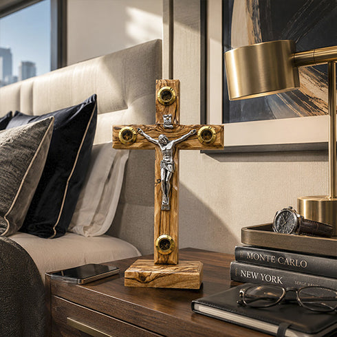 Wooden cross with gold accents on a nightstand next to a bed with pillows and a lamp.
