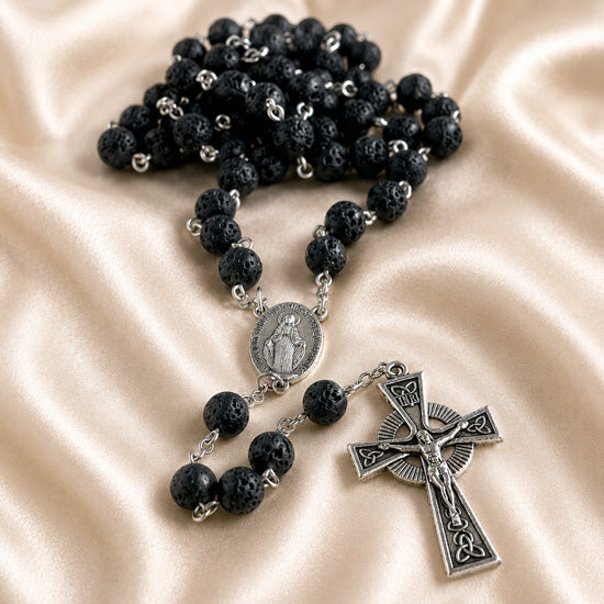 A black lava stone rosary with silver metal links, a round medallion, and a detailed crucifix pendant, on a satin background.