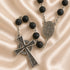 A rosary with black lava stone beads, a large ornate cross, a medallion displayed on a beige satin background.