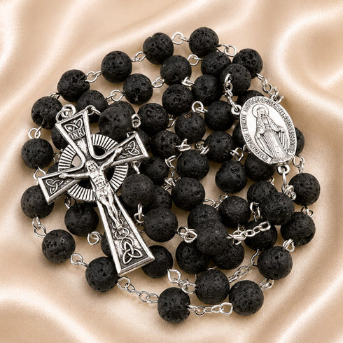 A black lava stone rosary with silver-tone metal links, a detailed crucifix, a round Miraculous Medal,on a beige fabric background.