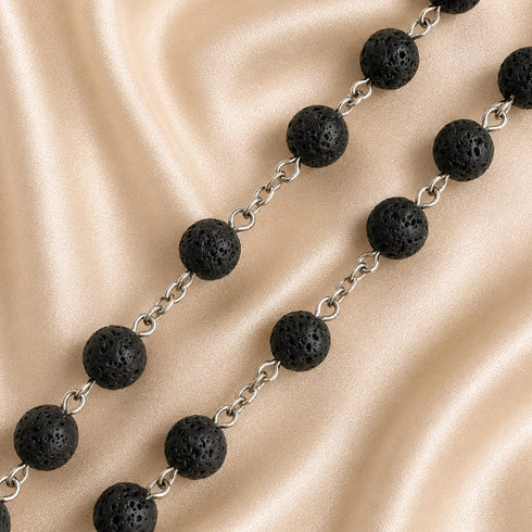 A strand of black lava stone beads connected by silver-toned metal chains, laid across draped satin fabric.