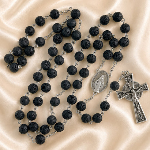 A black lava stone rosary with silver-tone chain links, a round medallion, and a crucifix, on a cream fabric background.