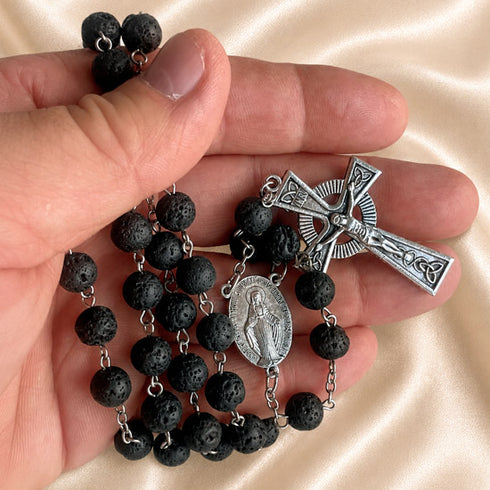A close-up of a hand holding a black lava stone rosary with a silver-tone crucifix and medal, set against a beige satin background.