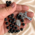 A close-up of a hand holding a black lava stone rosary with a silver-tone crucifix and medal, set against a beige satin background.