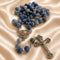A rosary with blue beads, a silver-tone crucifix, and a heart-shaped centerpiece with, on a satin fabric background.