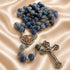 A rosary with blue beads, a silver-tone crucifix, and a heart-shaped centerpiece with, on a satin fabric background.