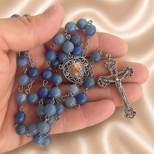 A rosary with blue marble beads, a silver-tone crucifix, and a central medallion, held in a person's hand on a satin background.
