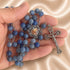 A rosary with blue marble beads, a silver-tone crucifix, and a central medallion, held in a person's hand on a satin background.