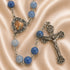 A rosary with blue beads, a silver centerpiece, and an ornate silver crucifix, on a satin beige fabric background.